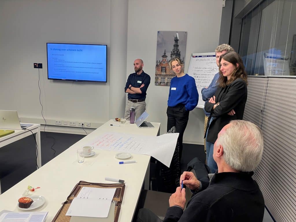 Vier personen staan voor een lokaal. Een man zit daarvoor aan een tafel waar pennen, papier en een kopje opstaat. Links van de groep mensen hangt een tv met een presentatie. 